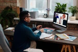 Man participating in a virtual meeting on a desktop computer with four colleagues visible on the screen, in a modern office setting with plants and natural light.
